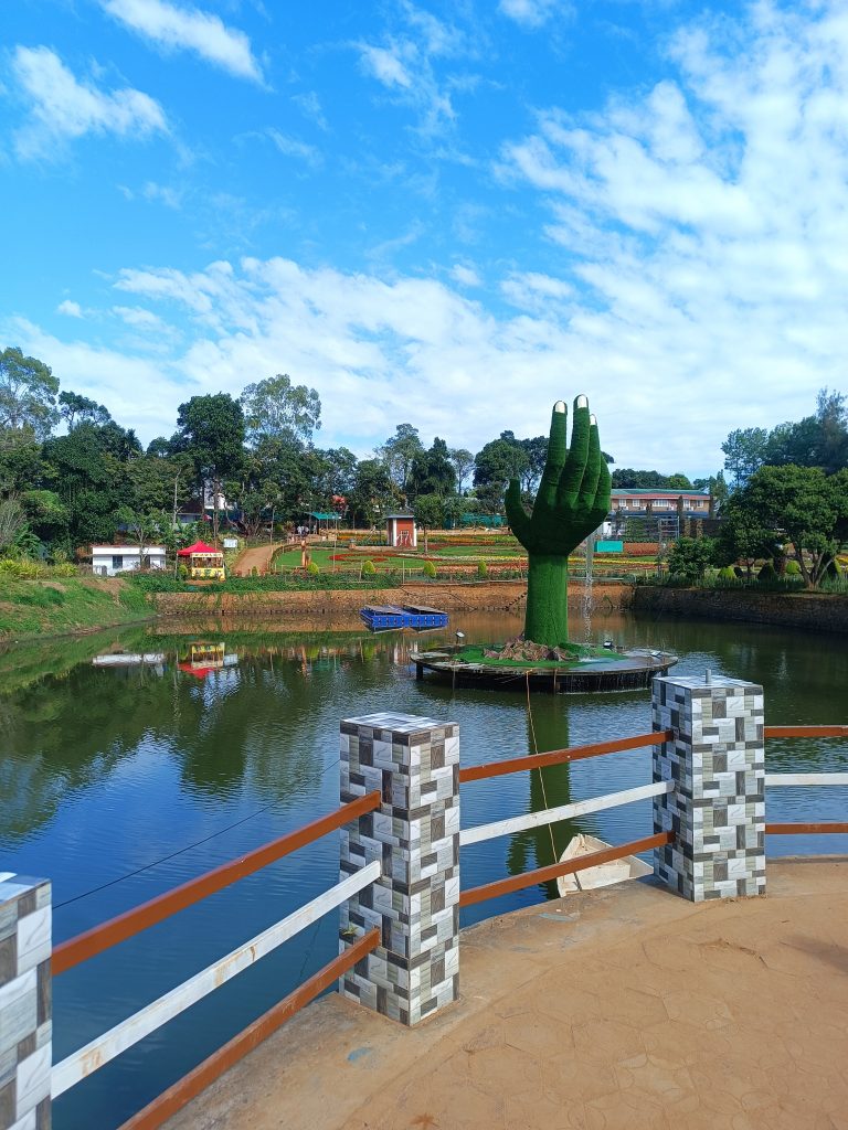 Park scene: Serene water surrounds a green hand statue, embraced by trees under clear skies.