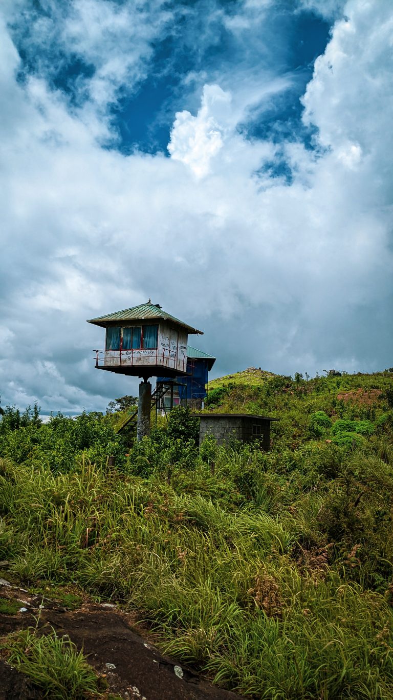 An elevated wooden watchtower with a sloped roof and graffiti stands on a lush green hill under a dynamic blue sky with white clouds. The structure is supported by metal poles above dense vegetation and a small concrete building.