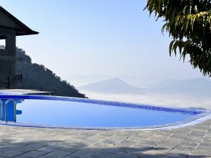 A tranquil swimming pool is situated upfront, and amidst the misty weather beyond, a large mountain is observable in Pokhara.