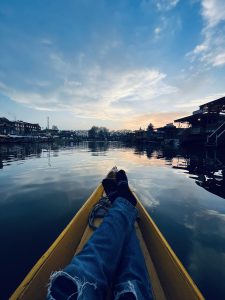 Person wearing a blue jeans reclining in a yellow boat on calm water at dusk, with silhouette of riverside houses against a cloudy sky, Kashmir Dale Lake.