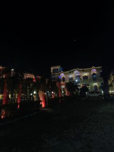 A building with lights all around illuminating it at night from every level. There is a car in front of the house and an outdoor patio lit up. 