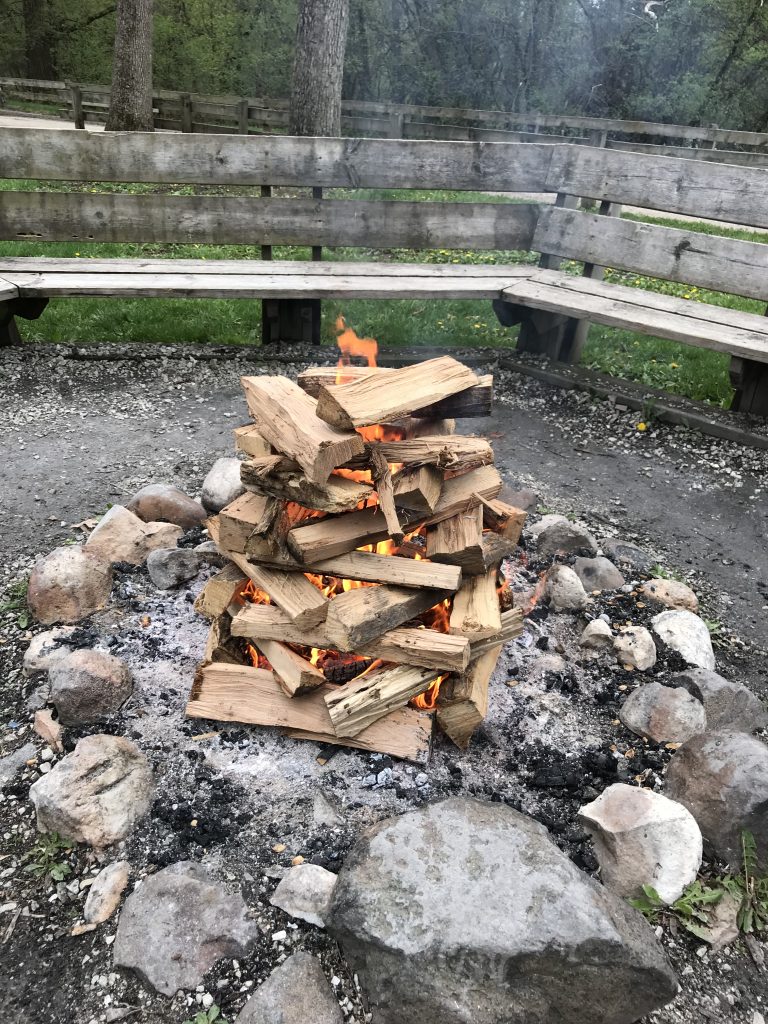 Fire ring with newly lit chimney-stacked firewood surrounded by wooden benches (YMCA Camp MacLean, Burlington, Wisconsin)