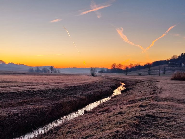 Sunrise in a winter in Central Europe (Slovenia) with a stream, fields, hills and open sky.