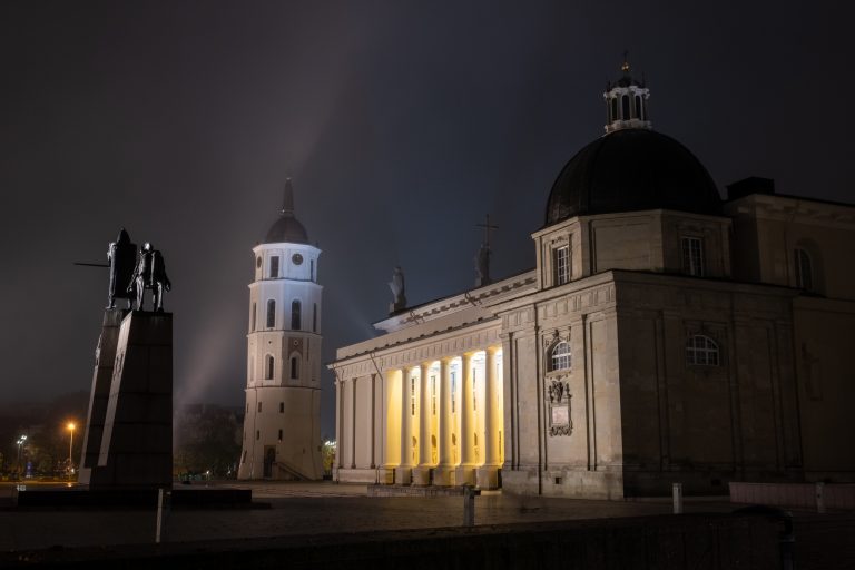 Nighttime photo of the Monument to Grand Duke Gediminas, bell tower, and Vilnius Cathedral with lights and a dark sky.