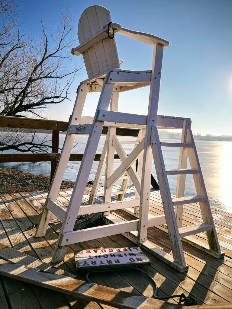 Lifeguard chair sitting on a beach dock at sunrise with a signing laying next to it describing “No Entry Without Lifeguard” (Fish Lake, YMCA Camp Duncan, Ingleside, Illinois)