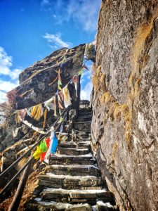 A narrow path goes up a mountain with a bit of snow on the steps. Big rocks are on each side, and colorful prayer flags wave along the path. 