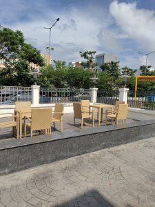 Brown tables and chairs in an outdoor patio beside a street. There are highrises in the background and a blue, cloudy sky overhead.