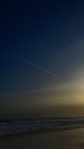 A jet's contrails illuminated on a dark night sky over waves on a beach