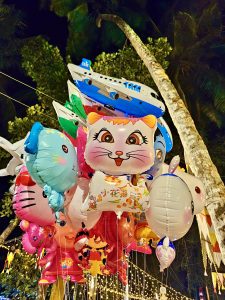 Balloons depicting bright, colourful cartoon characters, hang from strings ready for sale against foliage, the night sky and fairy lights. Temple festival, Kozhikode, Kerala. 