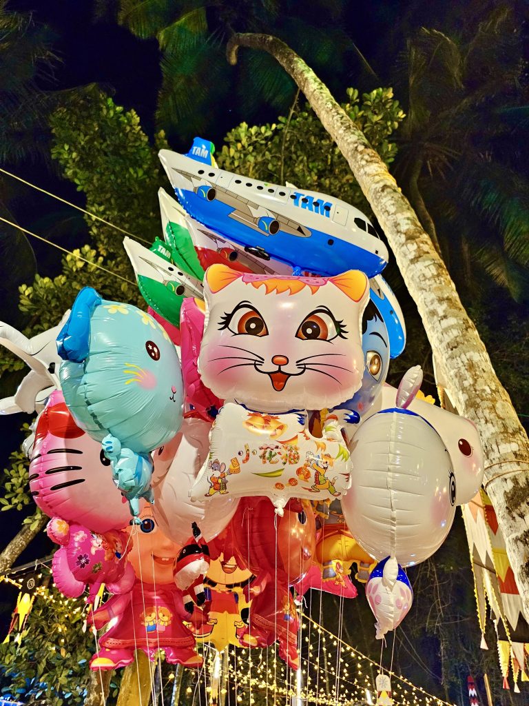 Balloons depicting bright, colourful cartoon characters, hang from strings ready for sale against foliage, the night sky and fairy lights. Temple festival, Kozhikode, Kerala.