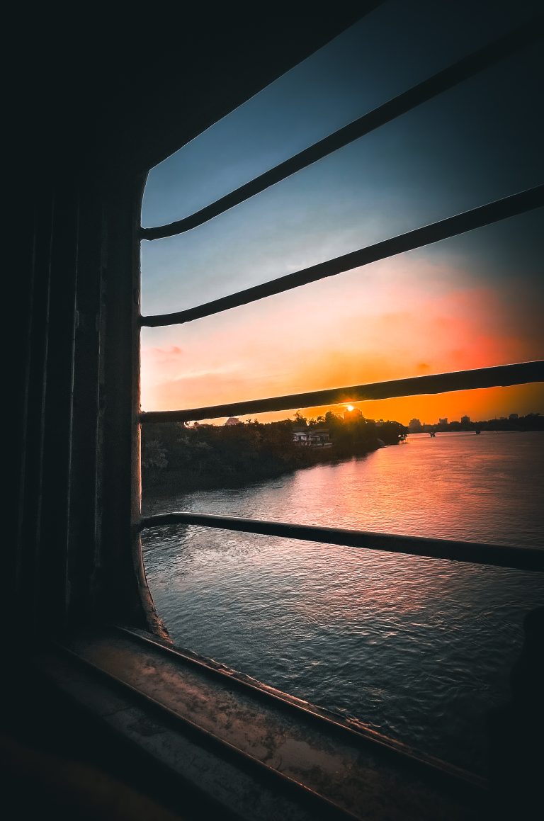 Sun setting behind the cityscape on the bank of a river, view from a train window.