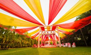 Colorful outdoor wedding stage (mandap) with radiant red and yellow fabric canopy, decorated stage, and chairs on lush green lawn.