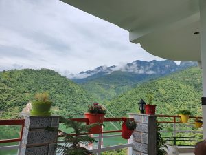 Vibrant greenery and a mountain range beneath clear skies, View from a balcony with colourful flower pots on the railings.
