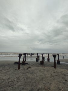 Seascape with remainder of Kozhikode bridge, only the rusted metal supports now visible protruding from the sea as the waves are rolling in to the beach. A couple of people standing on the shore. 