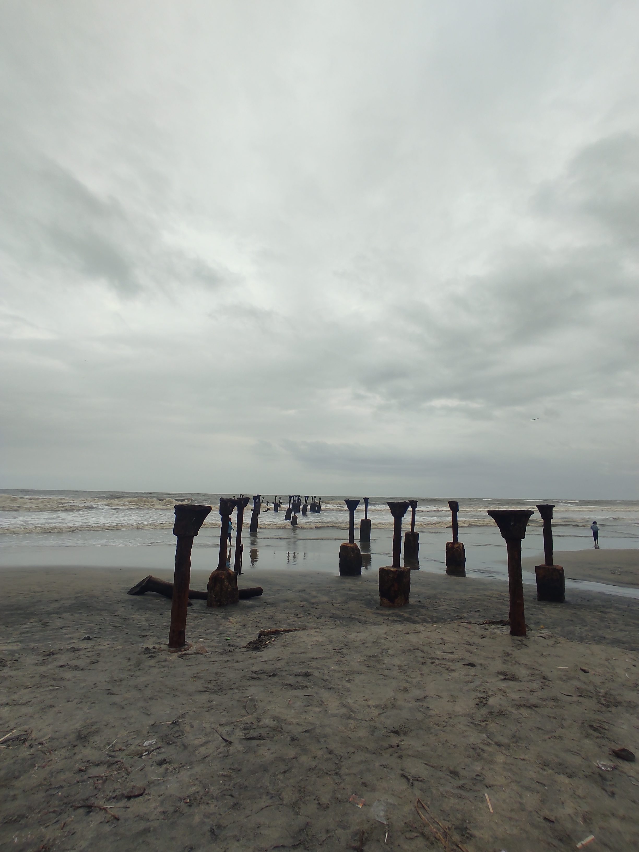 Seascape with remainder of Kozhikode bridge, only the rusted metal supports now visible protruding from the sea as the waves are rolling in to the beach. A couple of people standing on the shore.