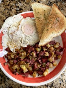 Homemade corned beef hash, two over-easy fried eggs, and two pieces of buttered toast are arranged on a white and orange plate, slightly overlapping each other. The plate sits on a brown and black granite countertop.
