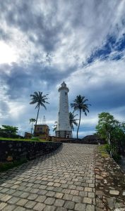 A white colored lighthouse with coconut trees near to it, with a background of cloudy blue sky. Taken from Galle fort, Sri Lanka.