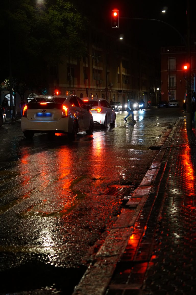 Rainy night in Sevilla, Spain. Cars driving down a city road are stopped as a pedestrian crosses the road.