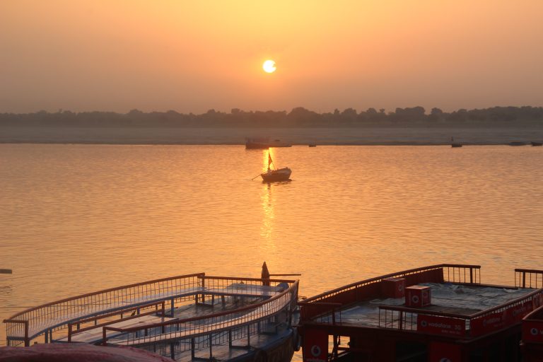 Sunrise view from a dock with some boats in the water and tree lines in the background. Dashassamedh Ghat, Benaras.