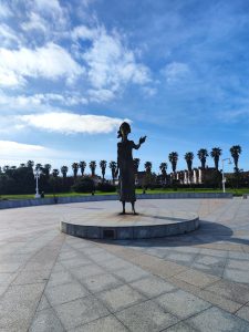 Silhouette of a statue (La madre del emigrante “La Lloca”) against a blue sky with scattered clouds, surrounded by palm trees in the background, on a circular platform ijón, Asturias, Spain
