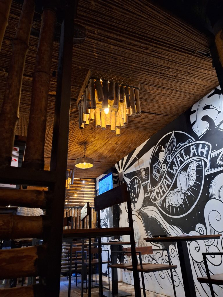 inside a chai shop: wooden tables and chairs. The ceiling is wood. There is a black and white mural on the wall that says “the chai walah.”