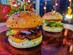 Chicken burger, garnished with cheese, red onion and lettuce. Buns sprinkled with black and white sesame seeds, presented ready to eat. 
