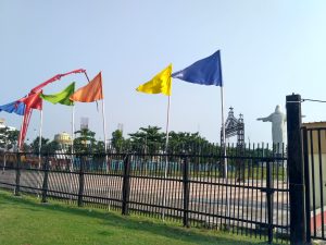 Colorful flags in a park behind a fence. There is a statue in the back to the right and an orange crane to the back left.