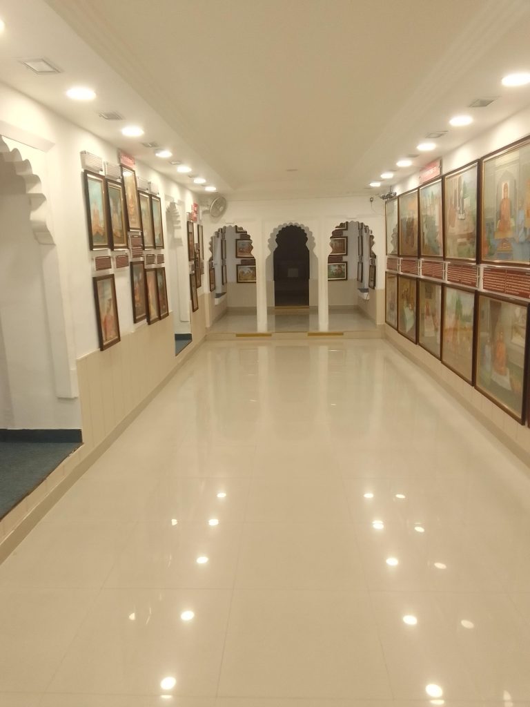 An art gallery with a long hallway and numerous framed paintings on both walls. The floor is glossy and reflective, and the ceiling has recessed lighting fixtures. An archway at the end of the hallway leads to another section of the gallery.