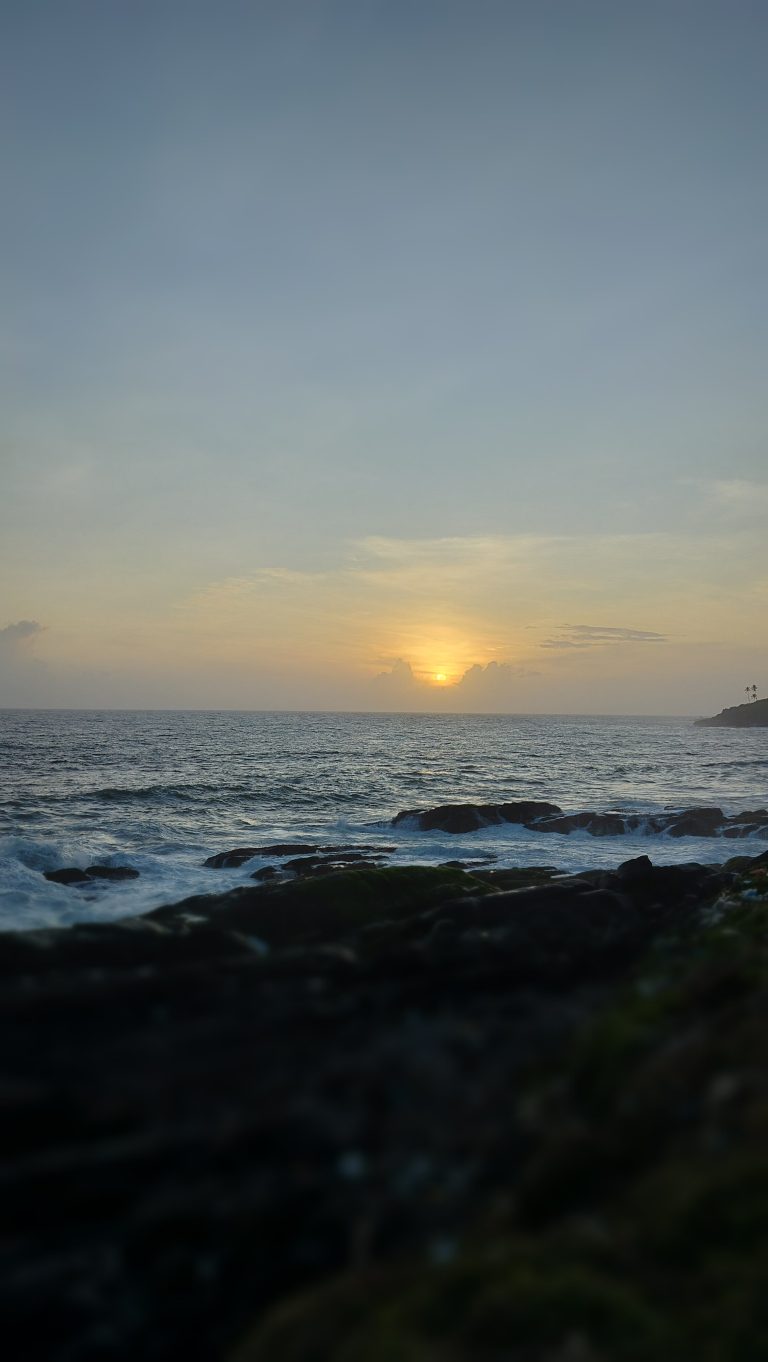 The sun low in the clouds over the ocean with waves crashing over large rocks at the shore