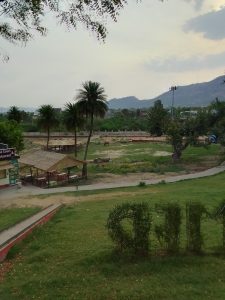 A serene park with lush greenery, palm trees, and a small building, set against a backdrop of distant mountains under a cloudy sky.
