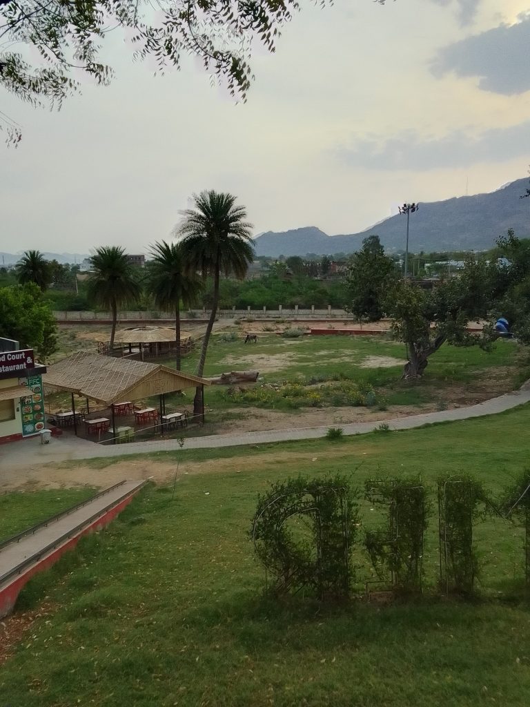 A serene park with lush greenery, palm trees, and a small building, set against a backdrop of distant mountains under a cloudy sky.