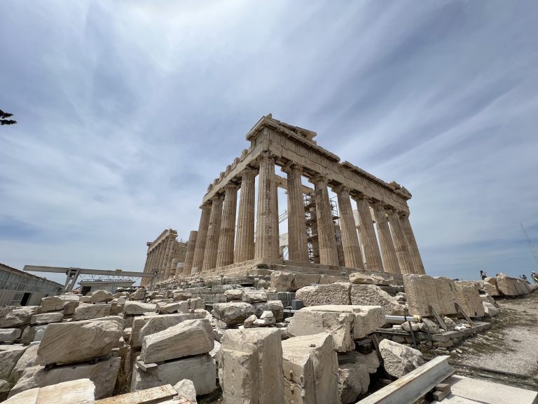 The Acropolis in Athens Greece. The sky is blue with some wispy clouds, and huge chunks of rock are visible in the foreground from ongoing maintenance and restoration efforts.