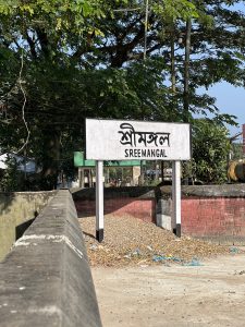 A signboard with the name “SREEMANGAL” in both Bengali and English, standing on concrete pillars with a background of green foliage, in front of a concrete wall.
