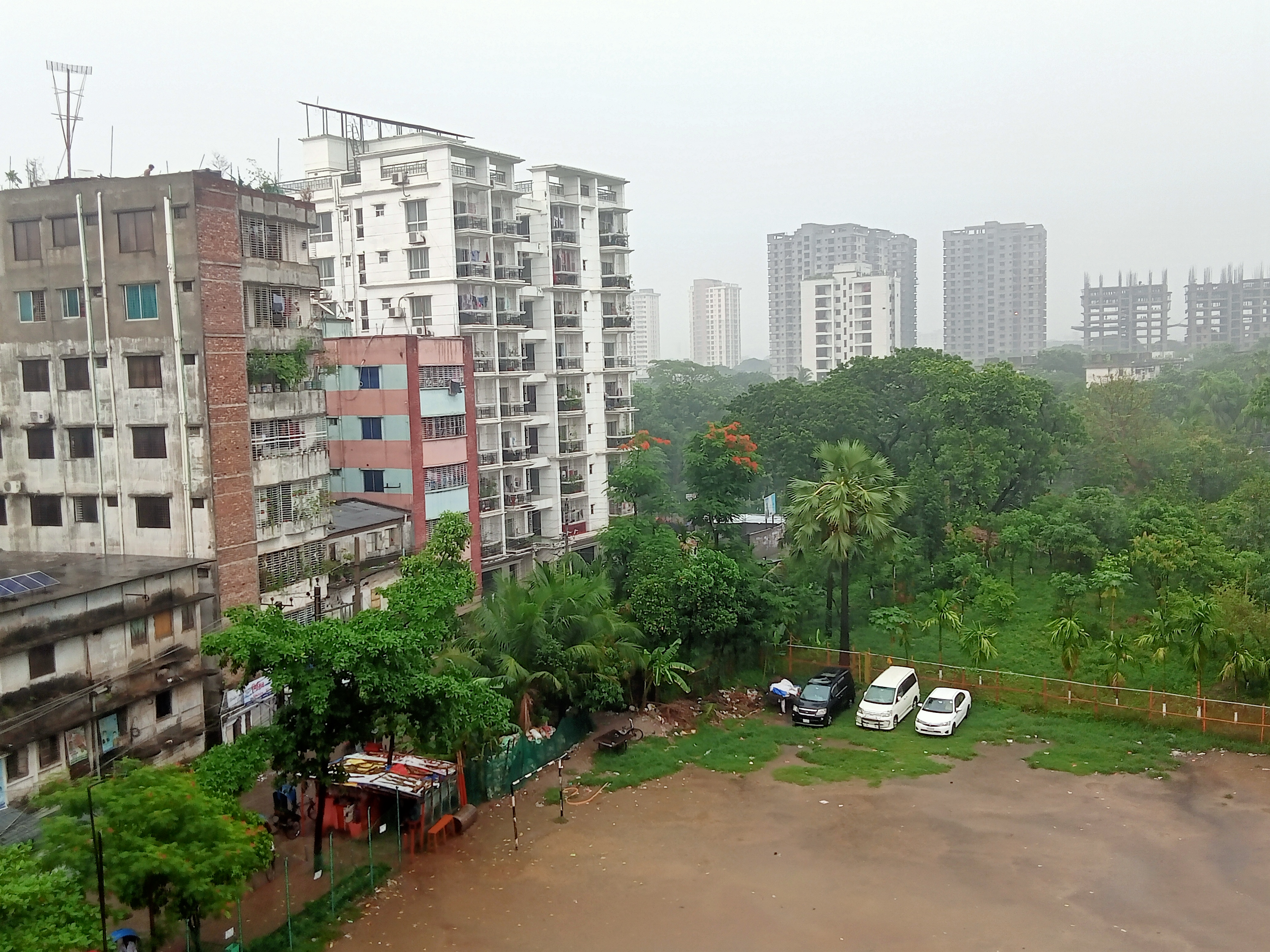 A rainy day in Dhaka, Bangladesh, with buildings beside trees and a dirt parking lot