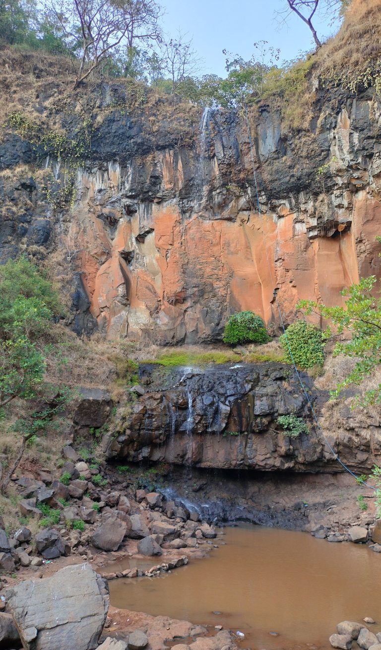 A waterfall with diminished flow over black and brown rocks into a pool below