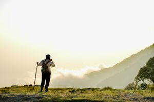 A man stands on mossy ground, carrying a rucksack on his back and bottle in his pocket, sips a drink while leaning on a walking cane as he takes in the mountain vista before him.