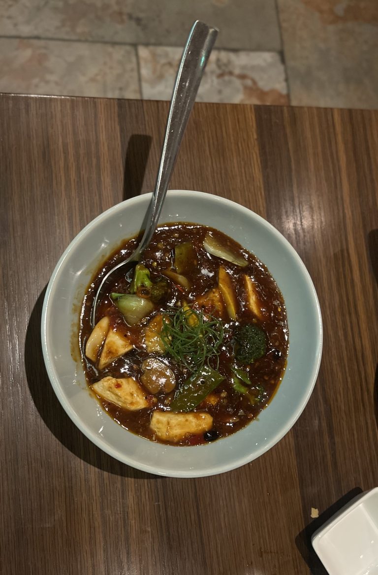 A bowl of spicy and savory Asian-style soup with tofu, vegetables, and a rich, dark broth, garnished with fresh herbs, served on a wooden table