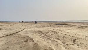 A sandy expanse at Alibag Beach near Mumbai; tire tracks crisscross, a quad bike and distant beachgoers under a hazy sky.
