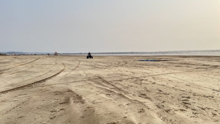 A sandy expanse at Alibag Beach near Mumbai; tire tracks crisscross, a quad bike and distant beachgoers under a hazy sky.