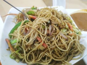 A plate of stir-fried noodles mixed with sliced vegetables such as bell peppers and onions, served with a side of soup.