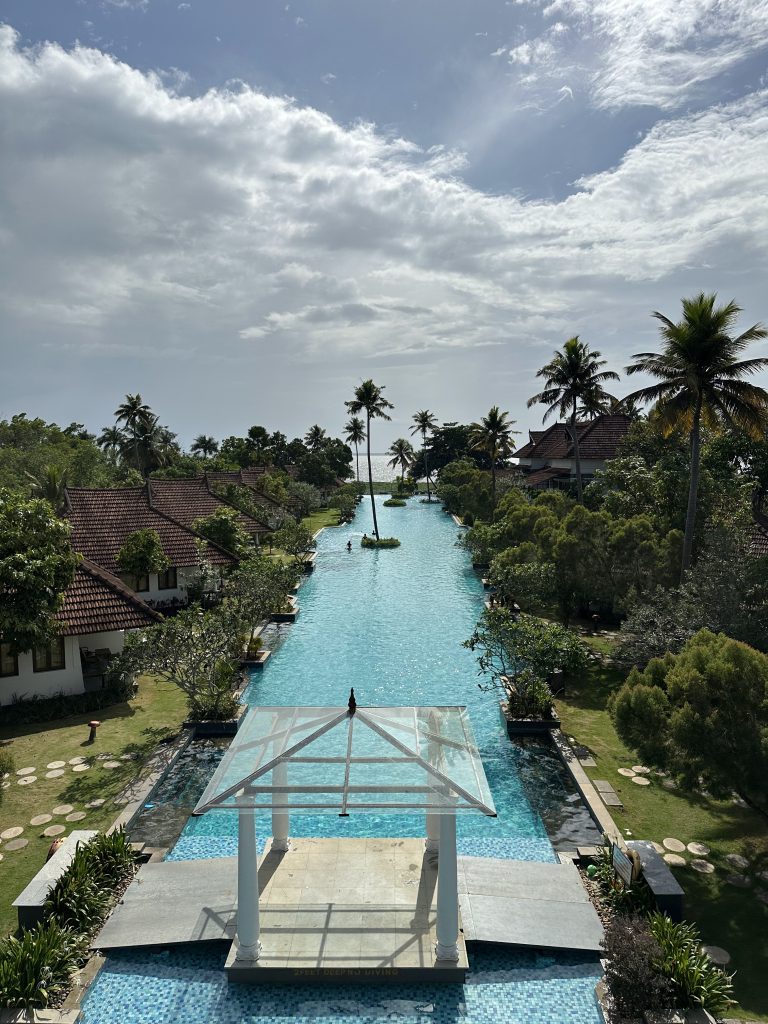 A sprawling pool, flanked by lush trees and buildings, with a distant lake adding to the serene horizon.