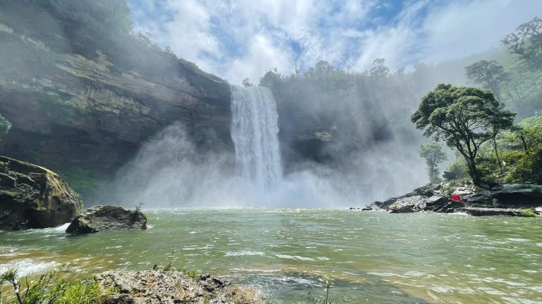 Waterfall pouring down a rocky cliff into a misty pool surrounded by lush greenery under a blue sky with clouds.