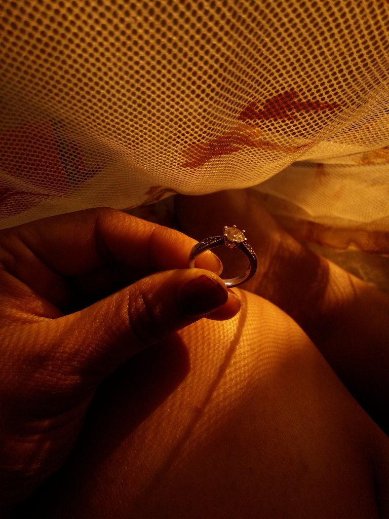 A hand holding a diamond ring illuminated by yellow light coming through a mesh fabric.