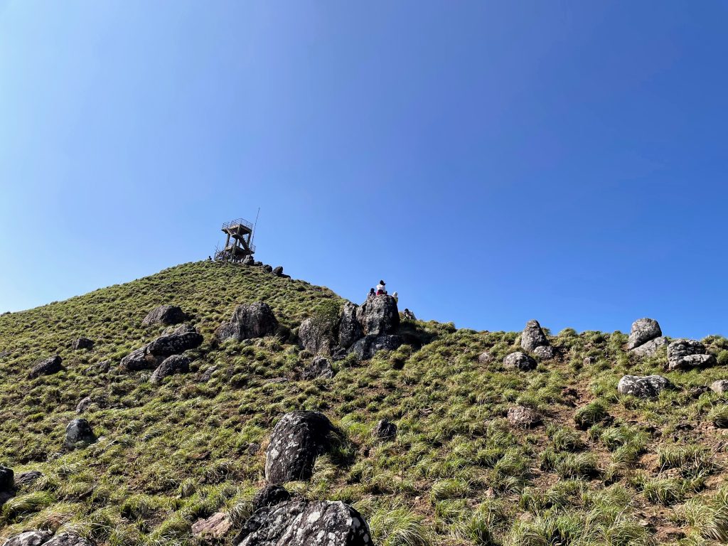 Looking up the mountain to Ponmudi, near Trivandrum, Kerala. It is a tower station at 1100m elevation. There is a bright blue sky, green grass, and people sitting on large rocks.