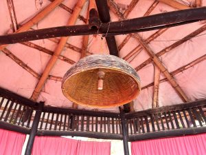 A light hanging under a Bamboo cane framed structure, paint peeling between the trusses. Red curtains hang below.
