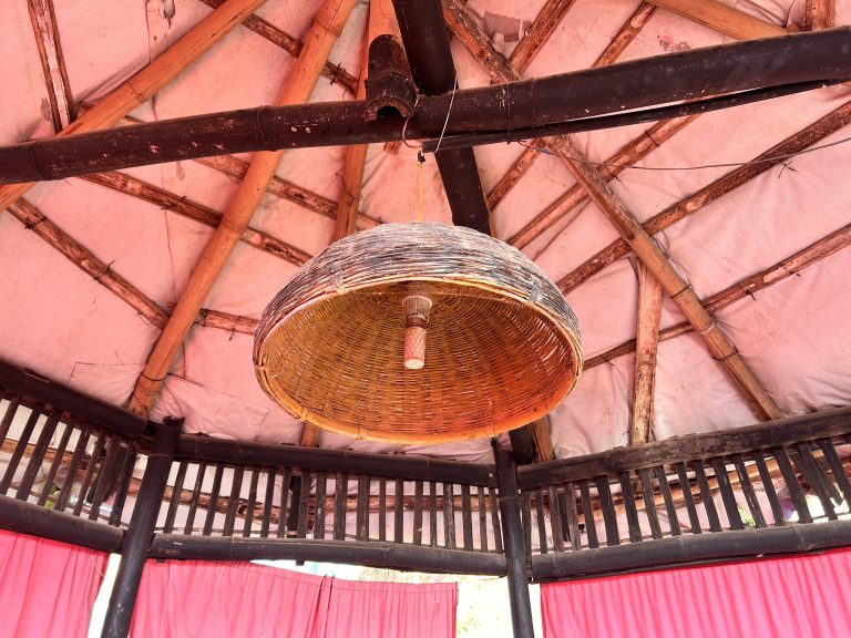 A light hanging under a Bamboo cane framed structure, paint peeling between the trusses. Red curtains hang below.