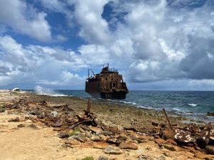 An oil tanker is stranded on the island of Klein Curaçao. The wreck is becoming increasingly rusty and less visible. The sea is filled with waves, and the sky is both blue and cloudy.
