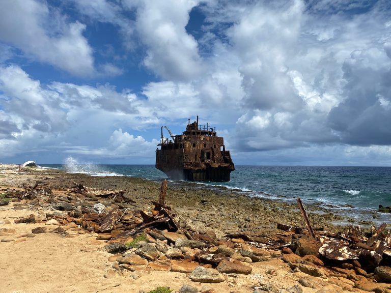 An oil tanker is stranded on the island of Klein Cura?ao. The wreck is becoming increasingly rusty and less visible. The sea is filled with waves, and the sky is both blue and cloudy.