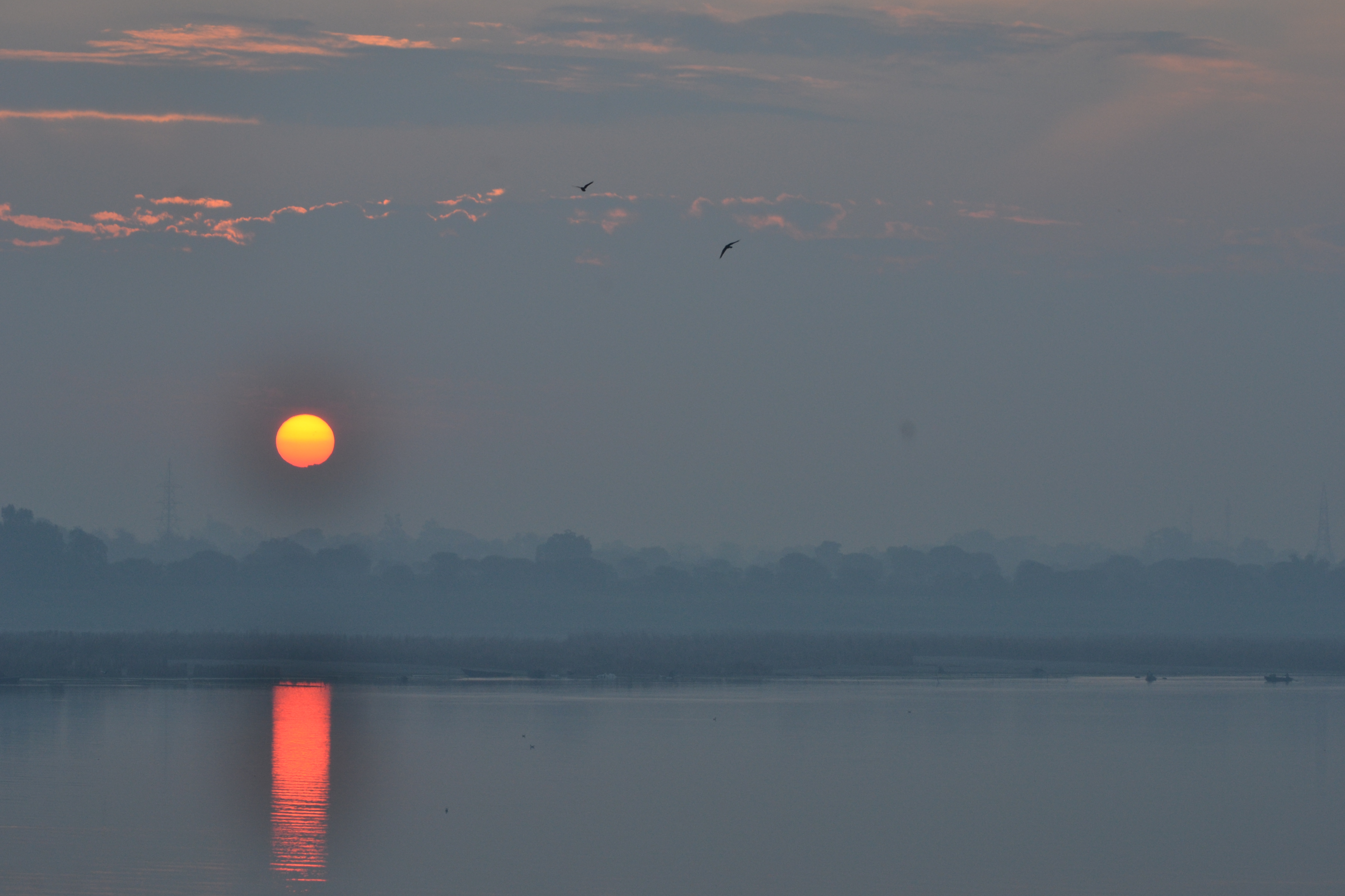 A mesmerizing sunset over the Ganga River, where the sky is painted with vibrant hues and the sun casts a reflection on the water. In the distance, birds soar homeward, completing the tranquil scene.