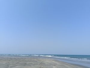 Beach with clear blue sky, gentle waves, and distant figures under the vastness of an open horizon.
